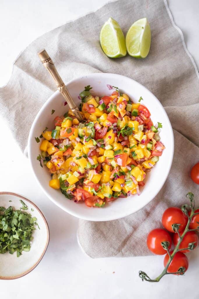 Mango cucumber salsa in a bowl, with a spoon in it. Tomatoes, coriander and lime wedges in the background